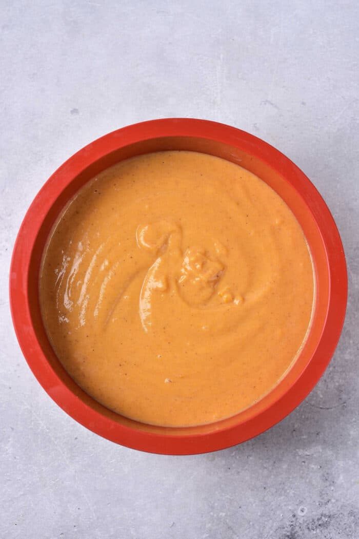 A red bowl filled with creamy, light brown Impossible Pumpkin Pie sauce sits on a gray, textured surface.