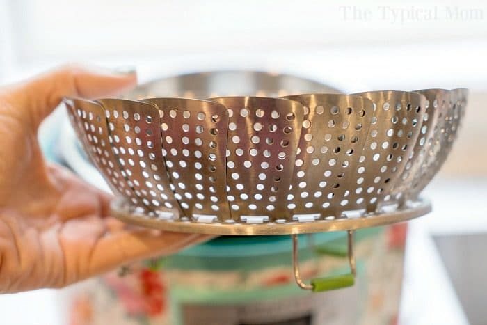 A hand holds a metal folding steamer basket over a bowl, showcasing its perforated texture and expandable design—an essential among instant pot accessories.