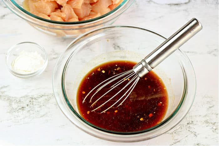Glass bowl with dark sauce and a whisk, solid for marinating asian chicken, sits next to a bowl of raw chicken and a small dish of white powder.