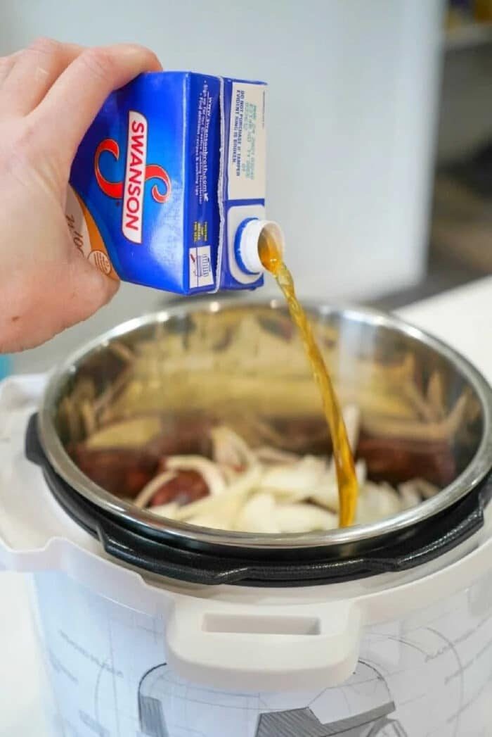 A hand pours Swanson broth from a carton into a pressure cooker filled with sliced onions, prepping for good instant pot BBQ beef.