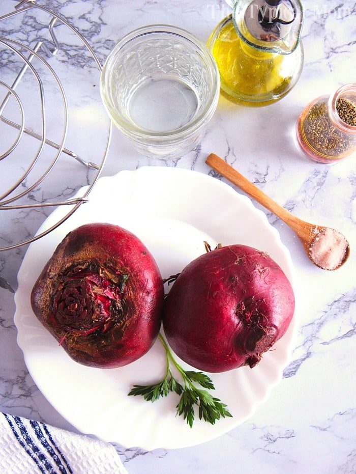Two whole instant pot beets rest on a plate with parsley, accompanied by a glass of water, oil, spices, and a spoon on the marble surface.