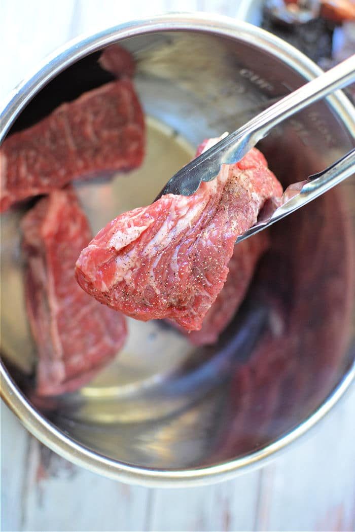 A pair of tongs holding raw beef over a metal pot, ready to transform them into flavorful Instant Pot Birria Tacos that are sure to delight.