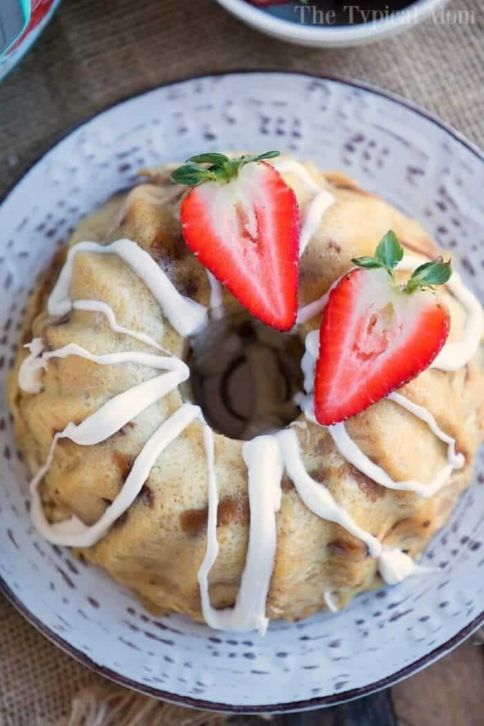 A bread pudding bundt cake with white icing, topped with halved strawberries, sits gracefully on a decorative plate, reminiscent of the cozy warmth that instant pot bread pudding brings to a home.