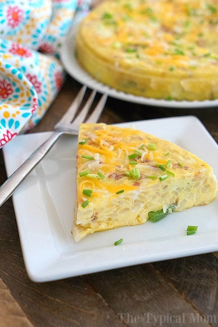 Slice of frittata garnished with chives and cheese on a square white plate, with a fork beside it. A full instant pot breakfast casserole graces a round plate in the background, next to a colorful cloth.