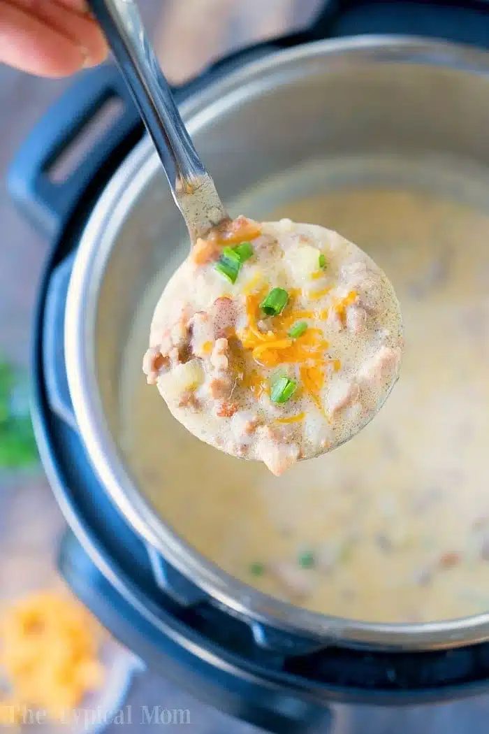 A ladle filled with creamy, instant pot cheeseburger soup brimming with chopped green onions, shredded cheese, and small meat chunks hovers above an Instant Pot filled with the same good soup. The edge of a cutting board showcasing extra toppings peeks in the background.