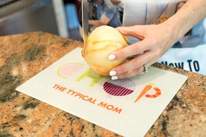 A person with white nail polish is slicing an onion on a cutting board marked with "CHOP Pynmi," preparing ingredients for an instant pot cheeseburger soup. The cutting board rests elegantly on a granite countertop.