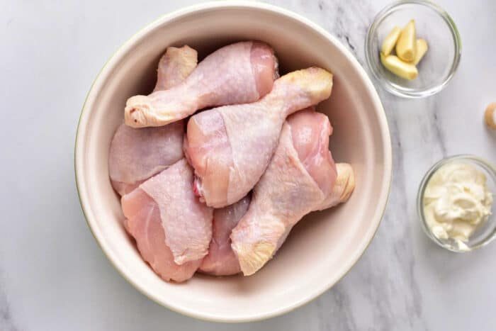 Raw chicken drumsticks in a bowl, with garlic cloves and mayonnaise in small bowls beside it on a white surface—solid for making flavorful instant pot chicken legs.