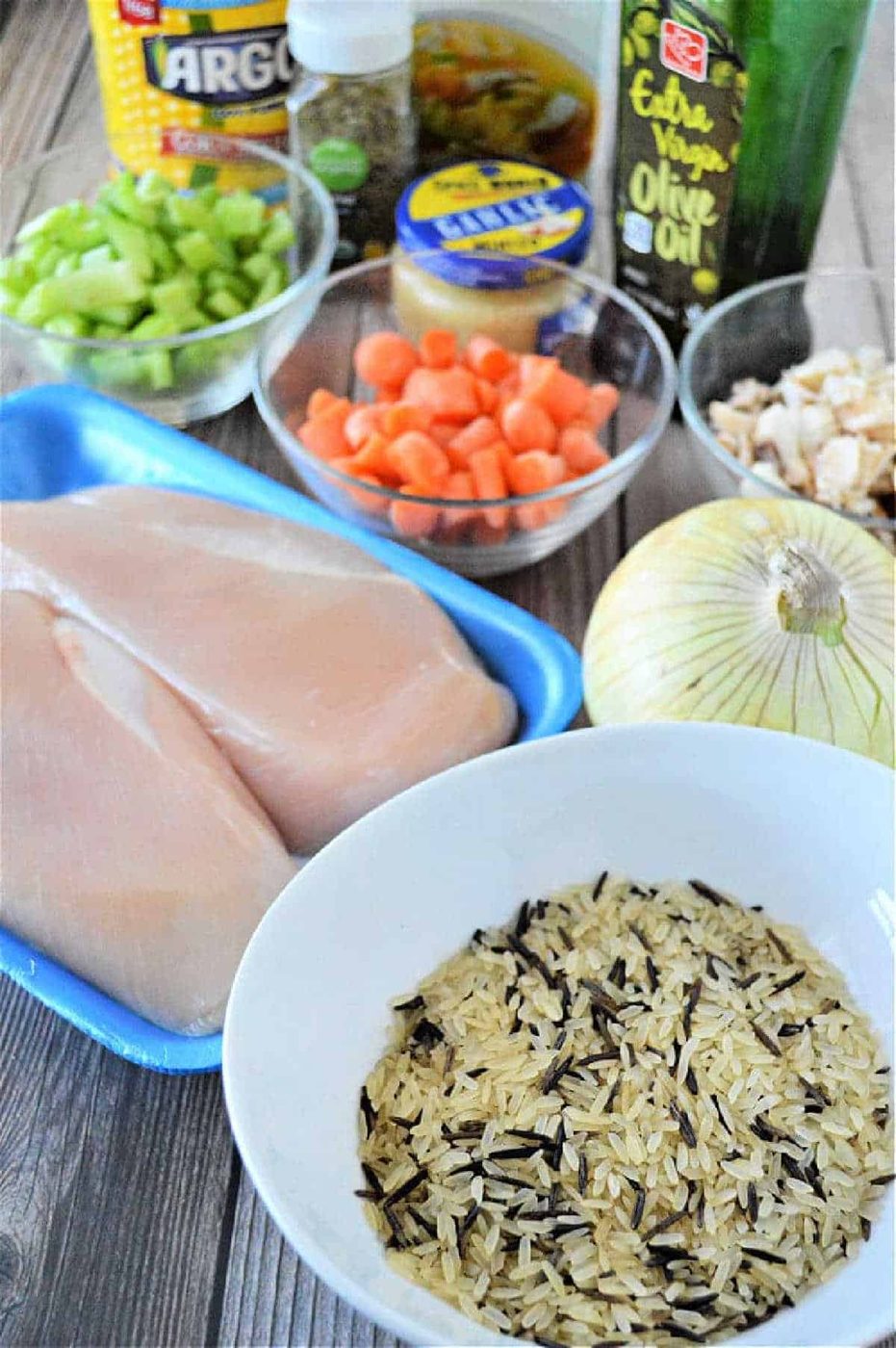 Raw chicken breasts, wild rice, chopped carrots, celery, onion, and various seasonings are ready on a wooden table—solid ingredients for a cozy instant pot chicken rice soup.