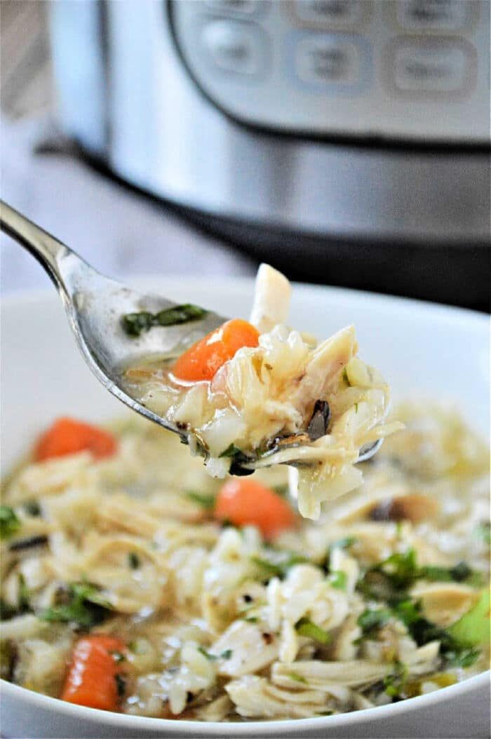 A spoonful of Instant Pot chicken rice soup is held above a white bowl, with an appliance in the background.
