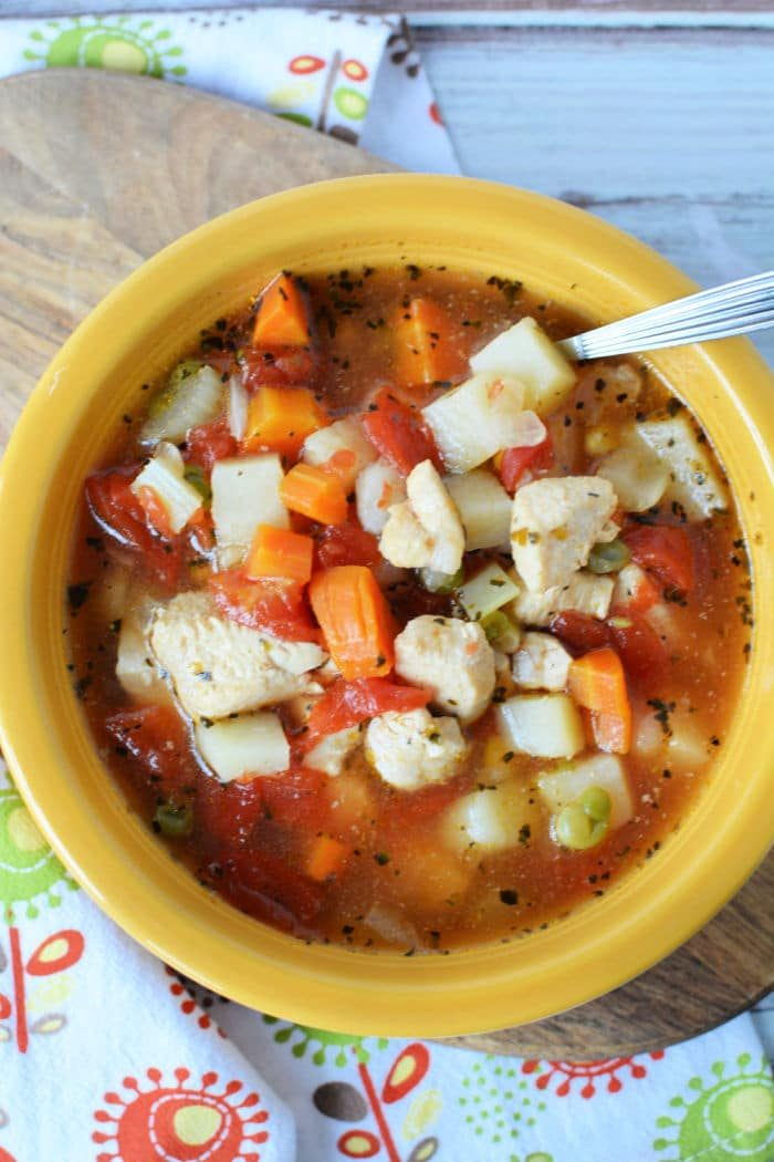 A yellow bowl of Instant Pot chicken vegetable soup brimming with diced potatoes, carrots, tomatoes, and peas, with a spoon resting inside. The bowl is placed on a decorative cloth.