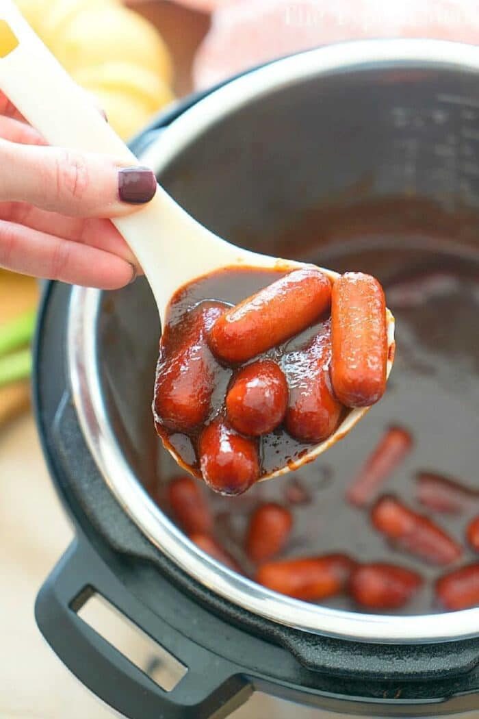A person holds a ladle filled with small sausages in a rich, brown sauce above an Instant Pot, perfecting the ultimate cocktail wiener recipe.