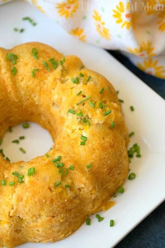 Golden cornbread ring on a white plate, garnished with chopped chives, reminiscent of an instant pot corn casserole recipe, with a floral napkin nearby.