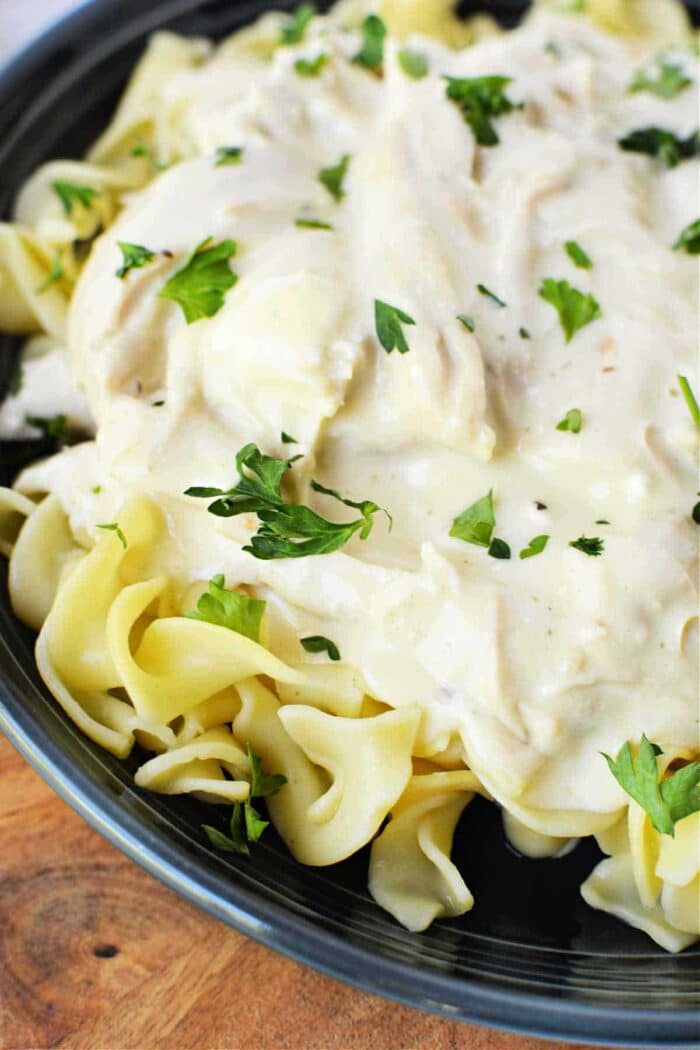 Instant Pot Creamy Chicken served over tender egg noodles, garnished with chopped parsley in a dark bowl.