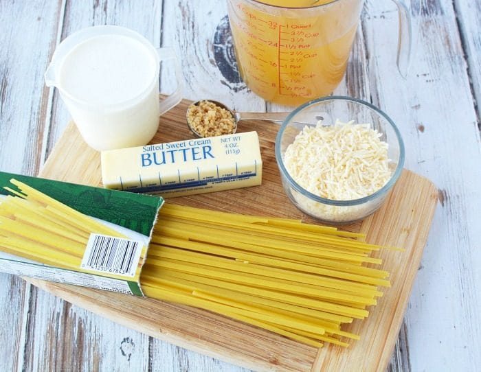 A wooden cutting board showcases uncooked spaghetti, a stick of salted butter, grated cheese, a measuring cup with liquid, a cup of cream, and a clove of garlic—solid ingredients for whipping up instant pot fettuccine Alfredo. The board rests on a rustic wooden surface.
