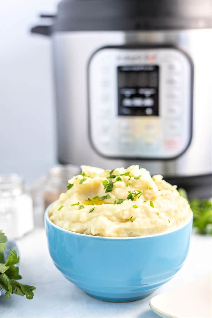 A bowl of creamy garlic mashed potatoes garnished with chopped herbs is in the foreground. In the background, there is an Instant Pot, its digital display glowing warmly on the kitchen counter.
