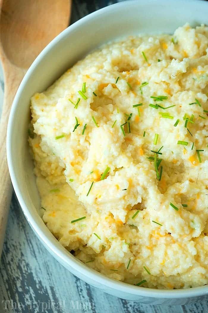 A bowl of creamy grits garnished with chopped chives sits invitingly alongside a wooden spoon. Nearby, Instant Pot grits offer a cozy pairing, solid for any comforting meal.