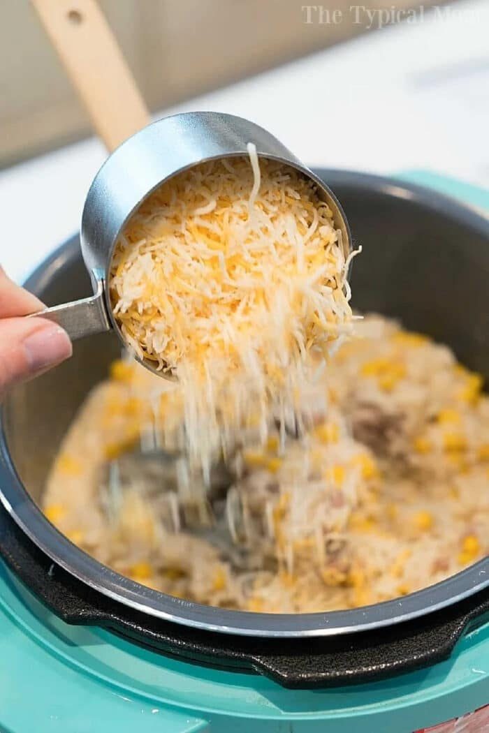 A hand pours shredded cheese from a measuring cup into a pot of rice and corn, like preparing an instant pot hamburger casserole.