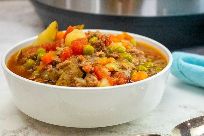 A white bowl filled with Instant Pot Hamburger Soup with Potatoes, peas, and carrots sits on a marble surface.