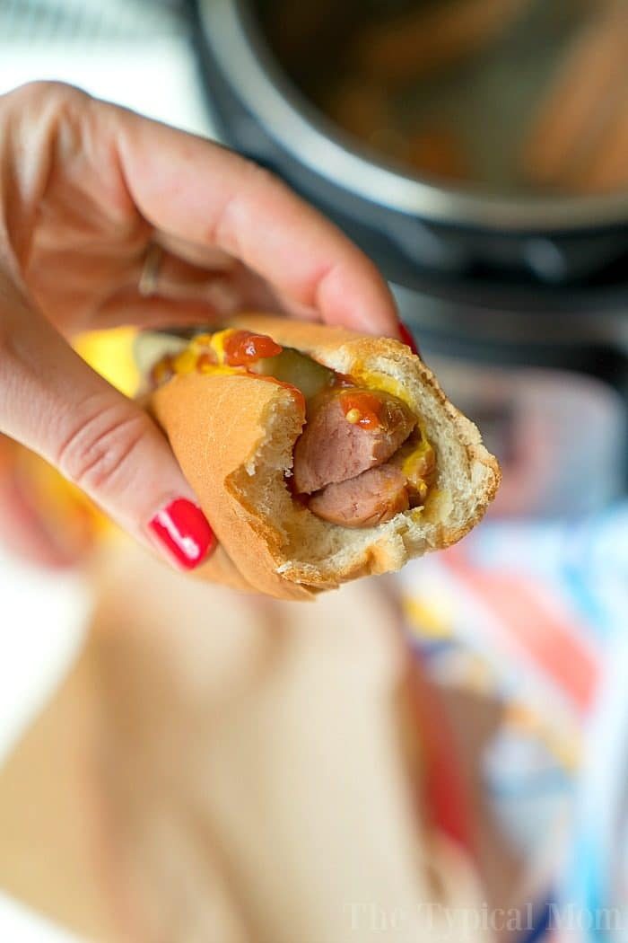 A hand holds a bitten hot dog with mustard and ketchup, the vibrant red nails adding a pop of color. Perfectly cooked using an Instant Pot, this hot dog promises both convenience and flavor in every bite.