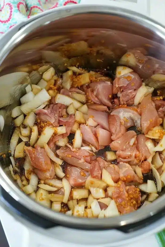 A metal pot contains uncooked diced chicken, chopped onions, and minced garlic. The ingredients are in the process of being sautéed, with some onions already starting to turn translucent, creating the base for a good Instant Pot Korean chicken dish.