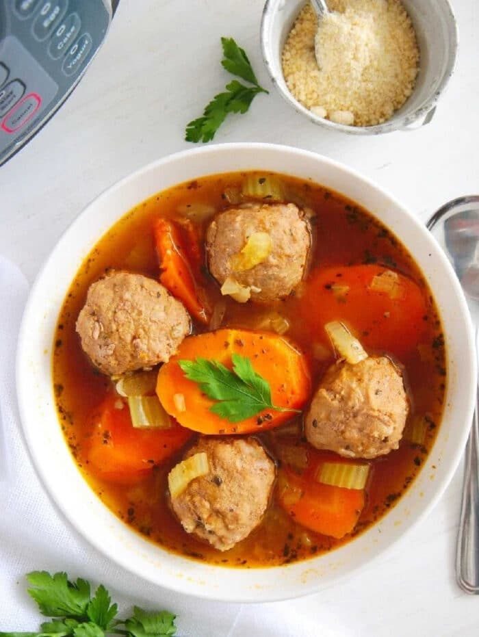 A bowl of Instant Pot meatball soup with carrots and celery, garnished with parsley, sits next to grated cheese and a spoon.