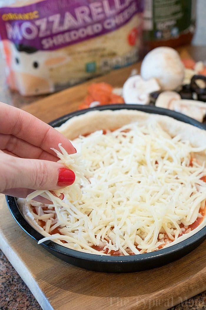 A hand sprinkles shredded mozzarella cheese onto a pizza crust with sauce, ready to be transformed into a good instant pot pizza. Mushrooms and olives nearby await their turn as tasty toppings.