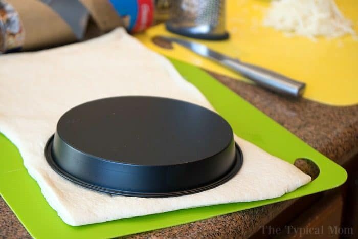 A baking pan is placed upside down on dough for instant pot pizza on a green cutting board with kitchen tools nearby.