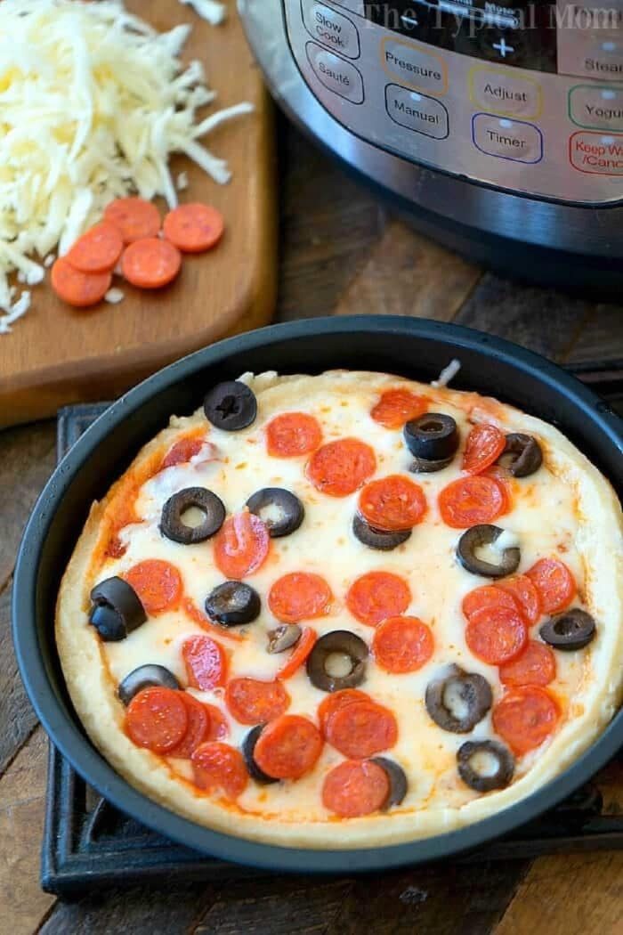 An instant pot pizza with cheese, pepperoni, and black olives sits near an Instant Pot and a cutting board with extra toppings.