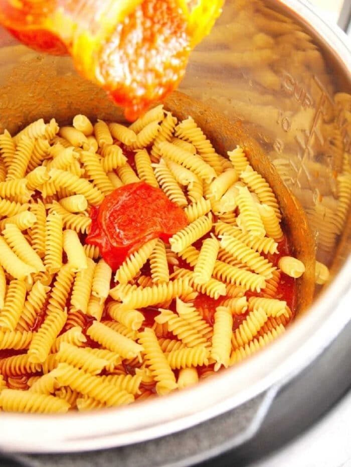 Rotini pasta and tomato sauce in a metal pot, with sauce being poured over the uncooked pasta—solid for making instant pot pizza pasta at home.