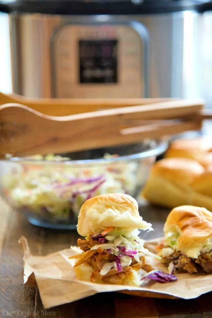 Pulled pork sliders topped with coleslaw on paper, with a bowl of slaw and a slow cooker in the background—solid for serving after making an instant pot pork shoulder recipe.