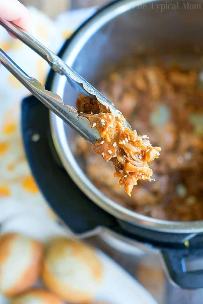 Tongs hold a portion of teriyaki pulled pork over an open Instant Pot. The succulent meat is mixed with sauce and sprinkled with sesame seeds. A blurred background reveals bread rolls and part of a printed cloth, adding a cozy touch to this culinary scene.