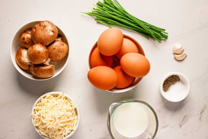 Brown eggs, mushrooms, shredded cheese, milk, chives, garlic, salt, and pepper arranged on a white surface—solid ingredients for a homemade quiche or an easy Instant Pot breakfast.
