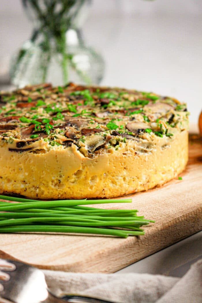 A thick vegetable frittata, inspired by an Instant Pot quiche recipe, is topped with herbs and sits on a wooden board next to fresh chives.