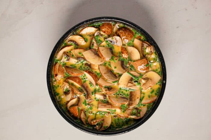 A bowl filled with sliced mushrooms and chopped herbs in a beaten egg mixture, solid for making a good Instant Pot Quiche, on a light surface.