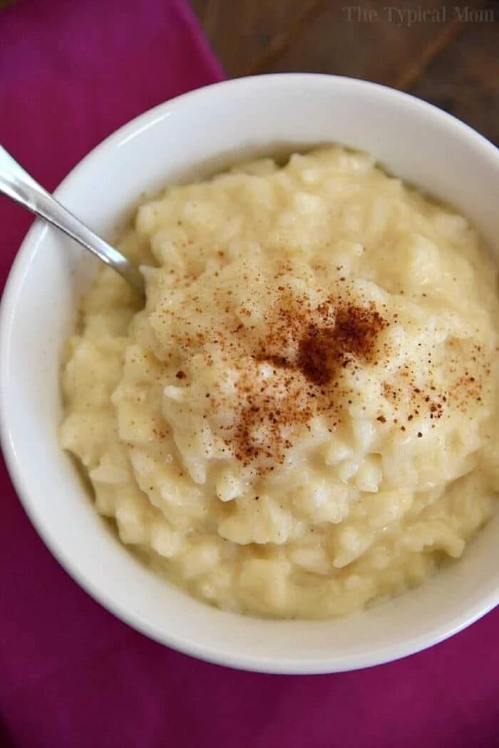 Bowl of creamy Instant Pot rice pudding topped with cinnamon, with a spoon, on a magenta cloth.
