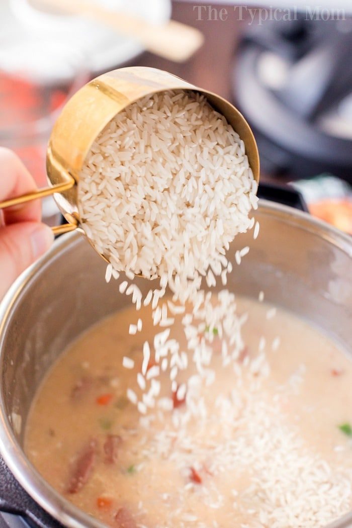 Pouring uncooked rice from a measuring cup into the instant pot of simmering sausage and rice casserole in pressure cooker