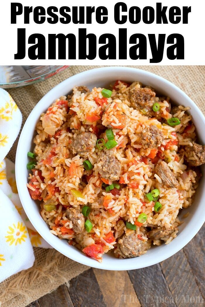 A bowl of savory jambalaya, crafted perfectly in a pressure cooker, sits on a wooden surface. It overflows with sausage, rice, tomatoes, and green onions. A white and yellow napkin rests beside this culinary delight.