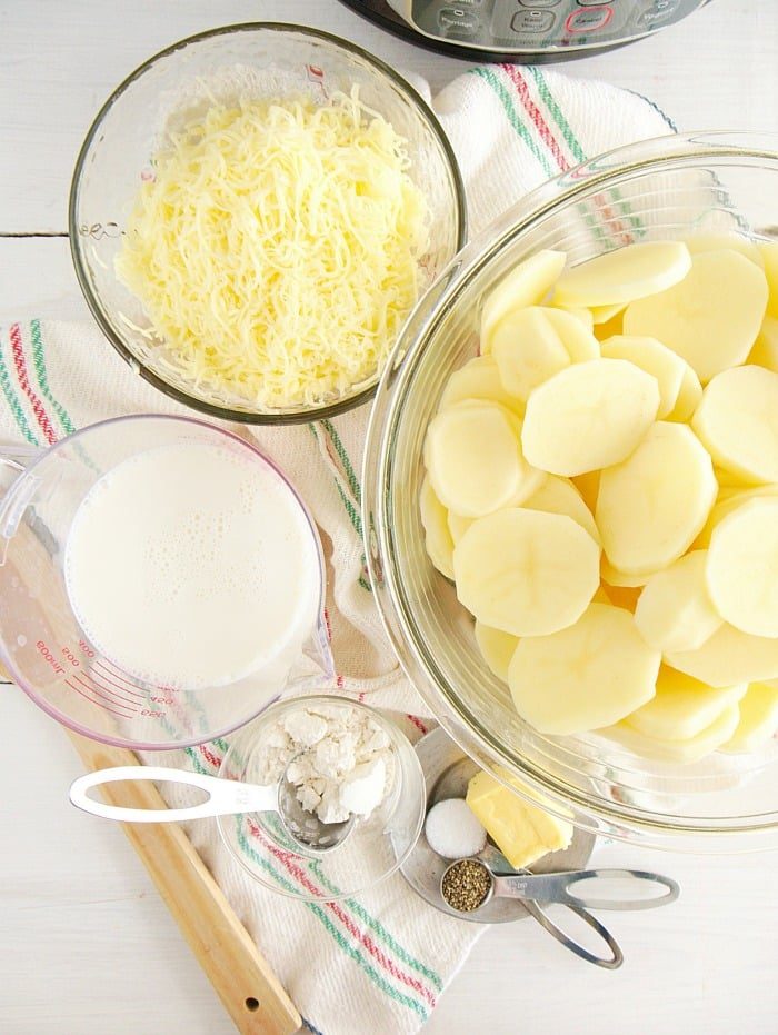 Ingredients for instant pot scalloped potatoes: sliced potatoes, shredded cheese, milk, flour, butter, and seasoning on a cloth.