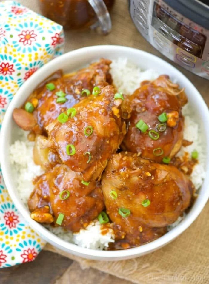 Bowl of saucy Instant Pot spicy teriyaki chicken thighs on white rice, garnished with chopped green onions, next to a pressure cooker.