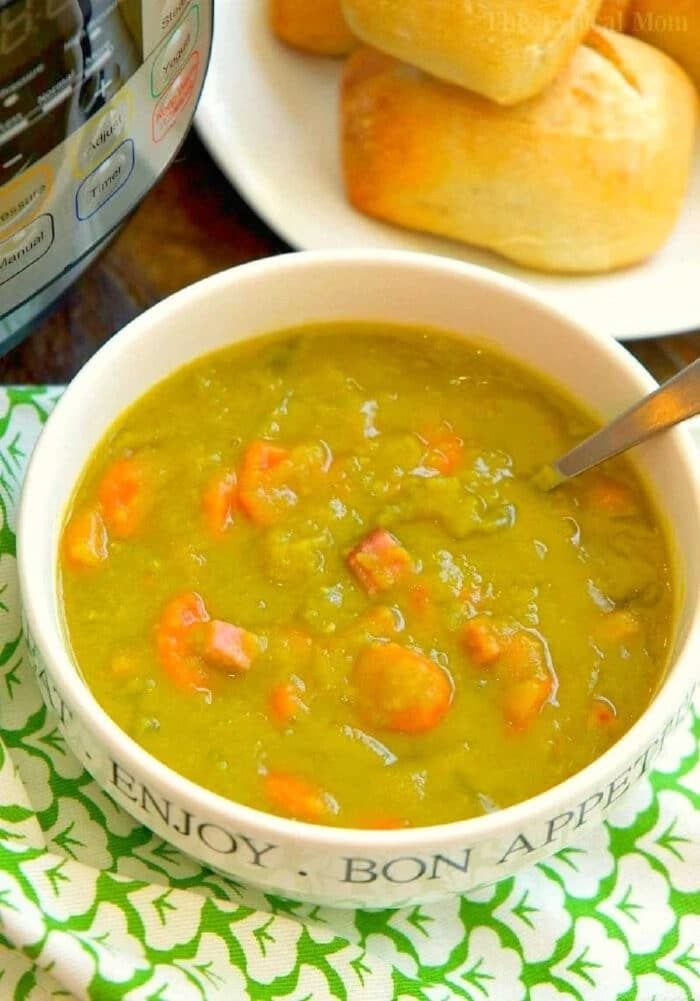 A bowl of Instant Pot Split Pea Soup Recipe with diced ham and carrots, served alongside a plate of bread rolls.