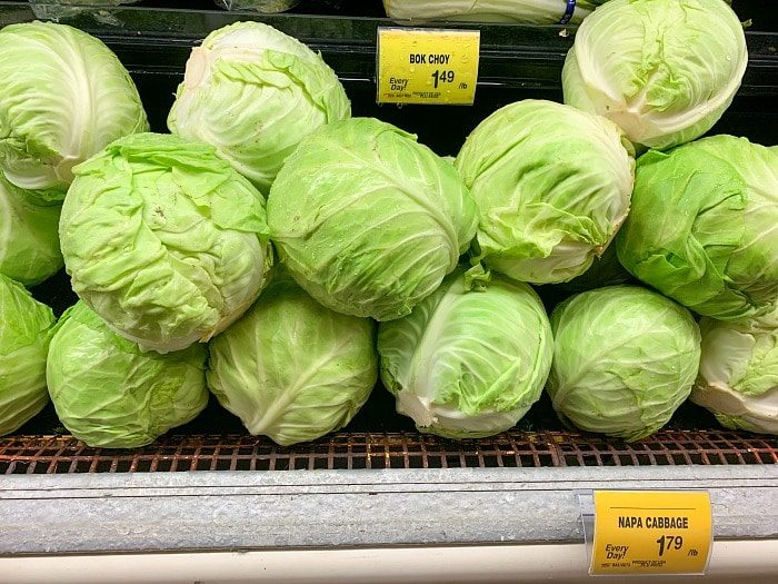 Heads of fresh green cabbage are neatly stacked in a grocery store display, accompanied by price tags for Bok Choy and Napa Cabbage.