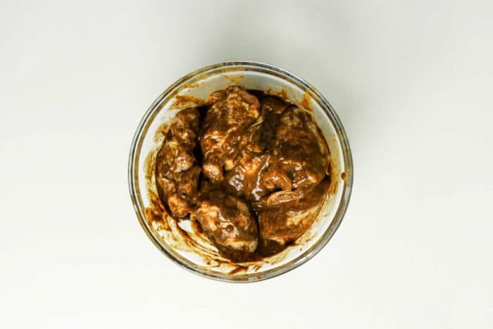 Glass bowl with pieces of meat coated in a thick brown jerk chicken marinade, ready for the oven, placed on a white surface—a solid start to your favorite jerk chicken recipe.
