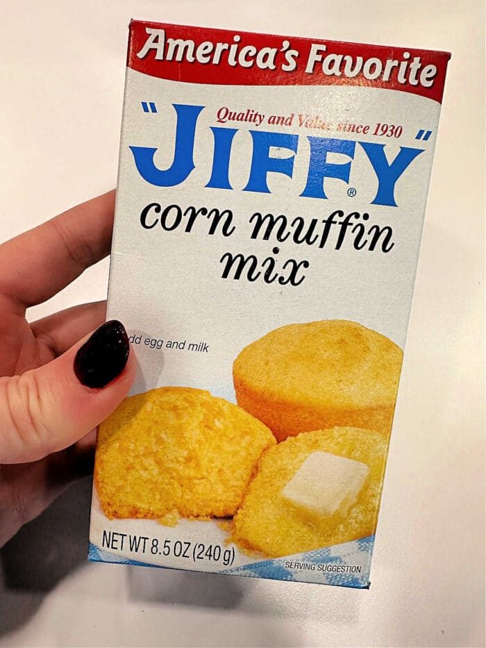 Hand holding a box of Jiffy Corn Muffins mix, featuring good muffins and a butter pat on the front—solid for trying your favorite corn muffin recipe or adding sour cream for extra moisture.