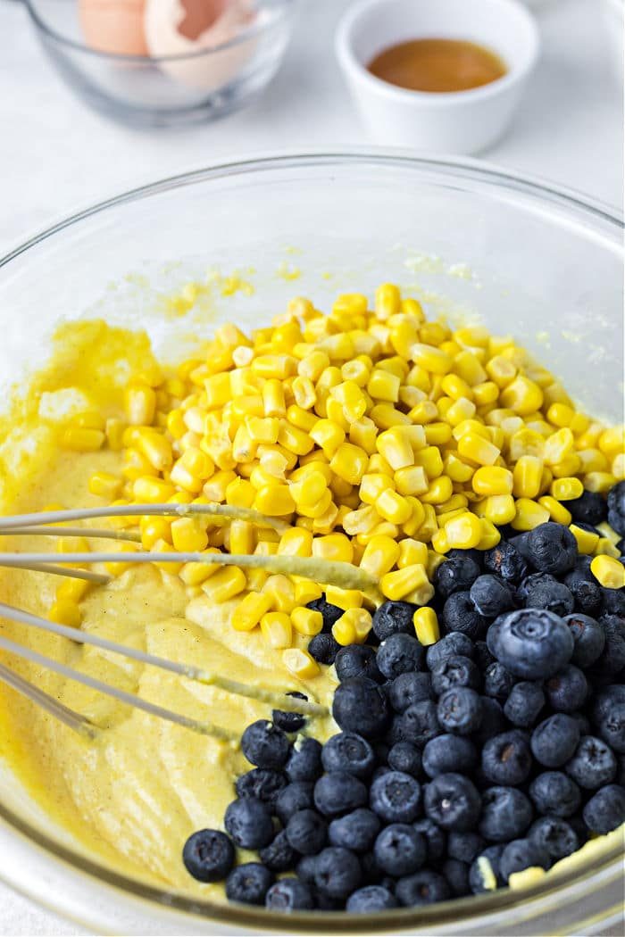 A glass bowl holds cornmeal batter adorned with corn and blueberries, whisk at the ready; eggshells and a cup linger in the background, setting the stage for a good jiffy cornbread baked to perfection in a cast iron skillet.