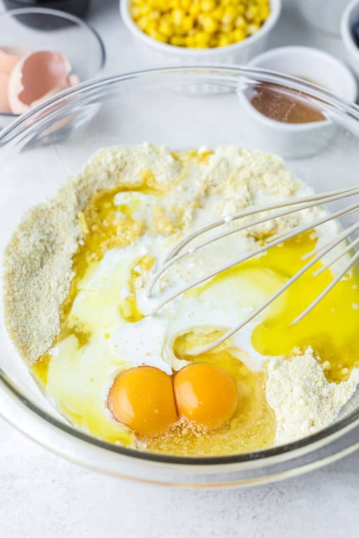 A mixing bowl with flour, two cracked eggs, and milk being whisked; corn and other ingredients in the background ready to create a good jiffy cornbread in a cast iron skillet.