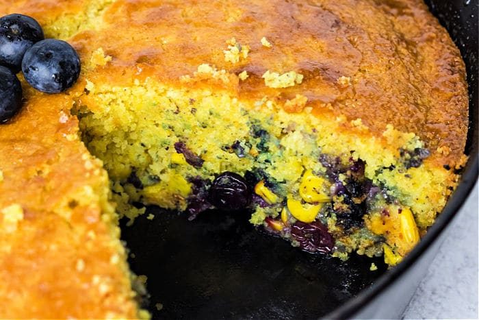 A close-up of blueberry cornbread made with Jiffy cornbread in a cast iron skillet, a slice removed to reveal its moist texture; topped with fresh blueberries.