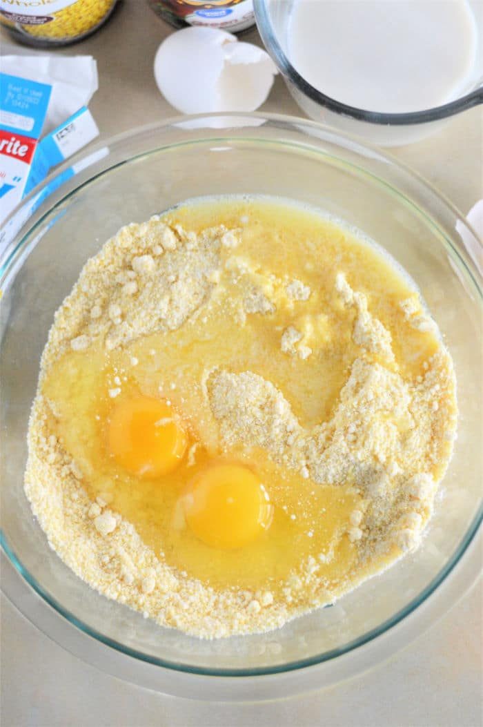 A glass bowl with Jiffy corn muffin mix, two eggs, and a liquid mixture sits ready to create Jiffy Mexican Cornbread, surrounded by a cracked egg and milk on the table.