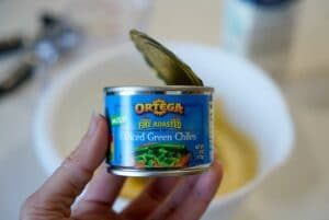 A hand holds a can of Ortega fire-roasted diced green chiles labeled as mild, solid for adding a kick to your dishes. The can is open, with one chile visible. In the background, you see a white bowl waiting to mix these chiles into an air fryer cornbread masterpiece.