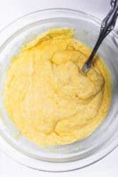 A glass bowl with yellow cornbread batter awaits a metal spoon, ready to transform this simple mixture into delightful corn dog bites.