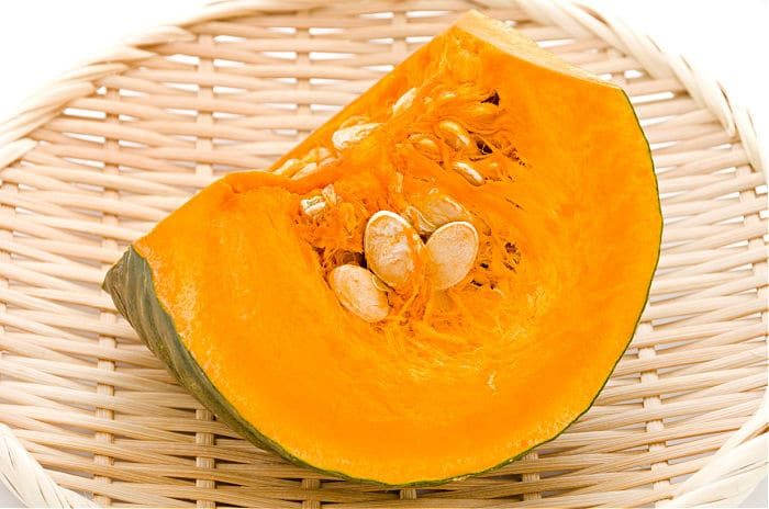 A slice of kabocha squash with seeds is placed on a round, woven bamboo plate, evoking thoughts of a comforting kabocha squash soup. The pumpkin's vibrant orange flesh and seeds are visible, with the rind near the outer edge against a crisp white background.